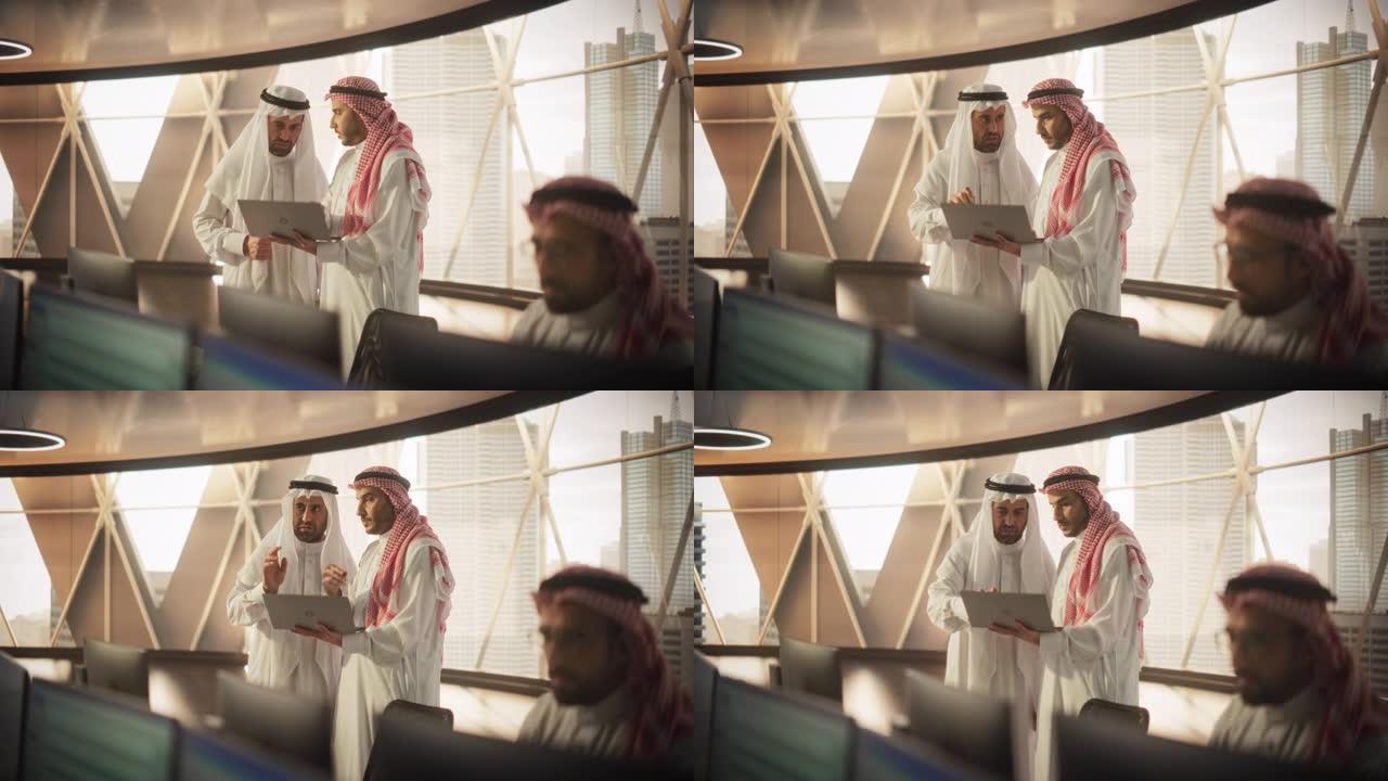 两名年轻的穆斯林财务顾问使用笔记本电脑在现代工业办公室讨论股票市场策略。阿拉伯经理人在银行研发中心工