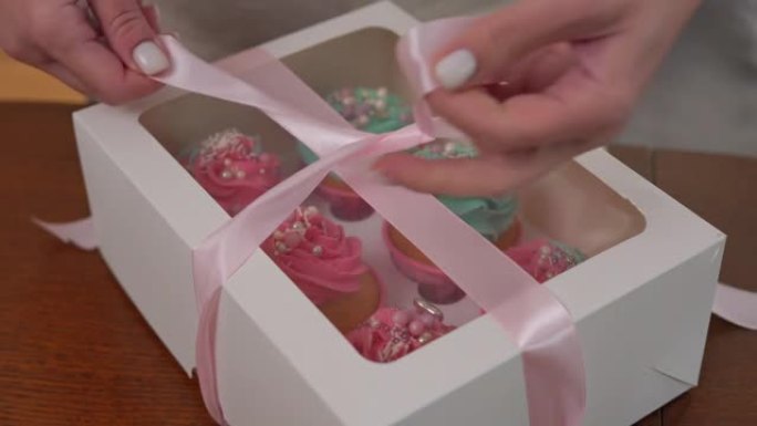 特写女性双手用松饼在纸盒上系上粉红丝带。面目全非的年轻白人妇女正在准备家庭面包店订单，以便在室内送货