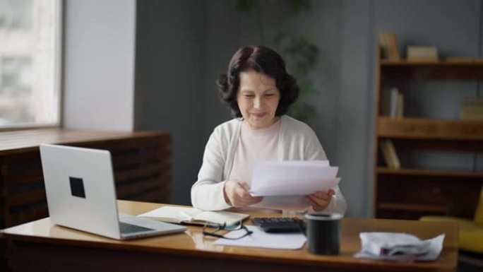 一位老年妇女坐在家里的办公桌旁，研究付款或税收。她检查文档中的数字，并将数据输入笔记本电脑。票据支付