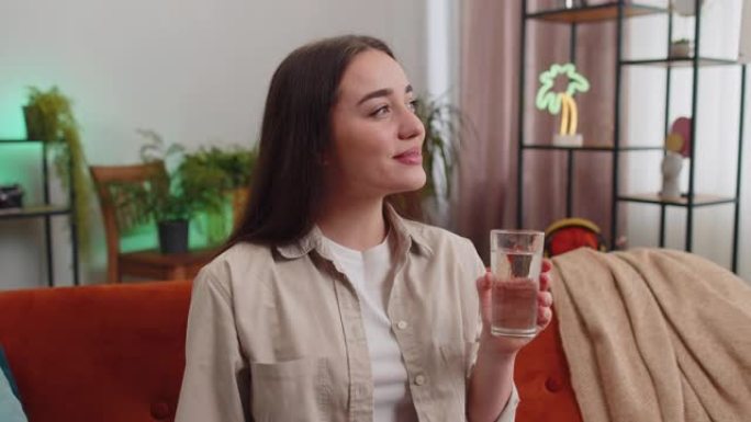 口渴的年轻女子坐在家里拿着一杯天然水，喝了口水
