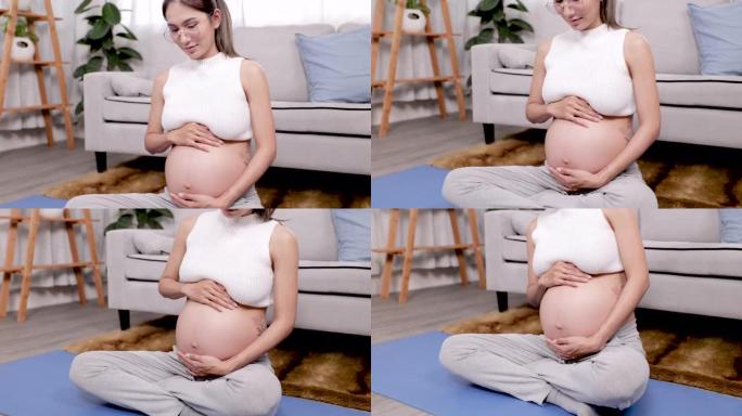 4K，美丽的亚洲孕妇，大肚子，坐在蓝色的运动垫上，她坐下来轻轻抚摸着成长中的肚子，刺激未出生的孩子，