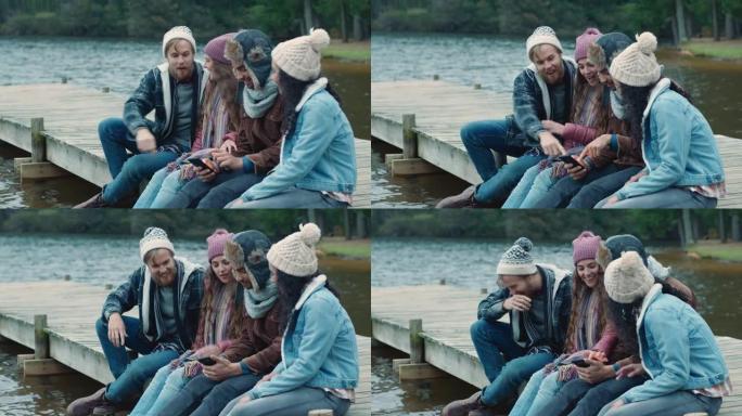 使用智能手机浏览照片的朋友一起分享回忆，坐在湖边的码头上聊天，在阴天的冬日穿着暖和的衣服闲逛