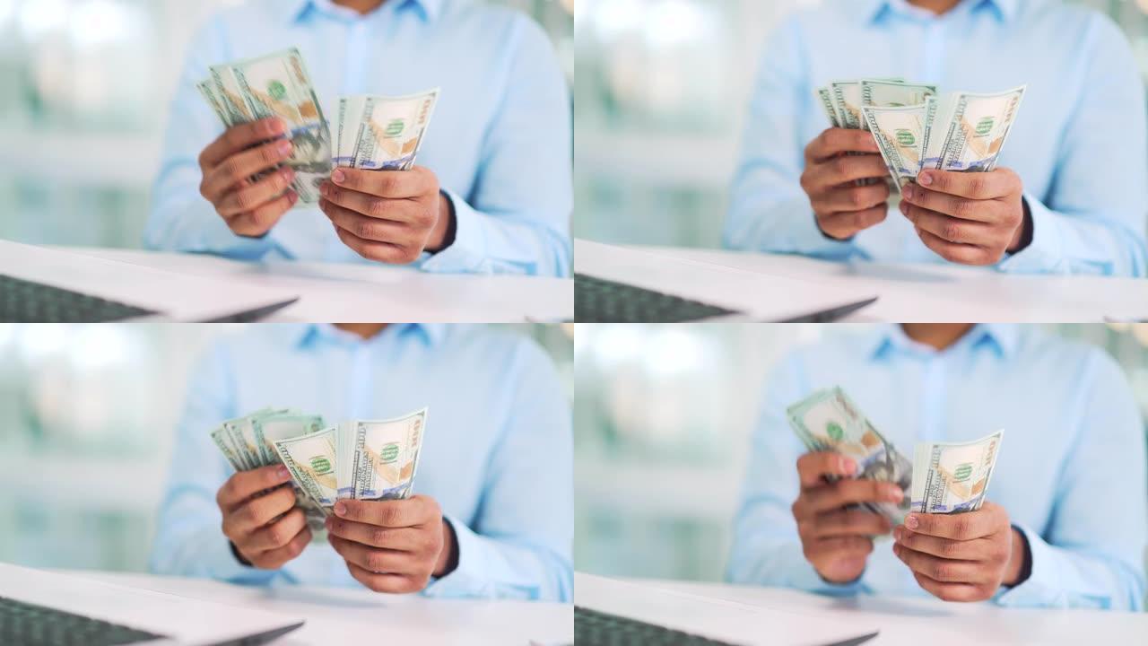 男性手数百美元钞票的特写。一位穿着衬衫的商人在现代办公室的工作场所的办公桌上数钞