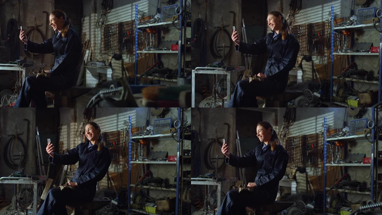 穿着制服工作服的年轻女机械师的肖像使用智能手机与家人和朋友进行视频通话聊天。在维修车间休息的焊工职业