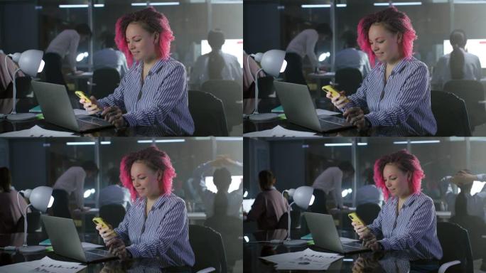 粉红色头发的办公室经理在笔记本电脑上工作，并在办公后期使用智能手机