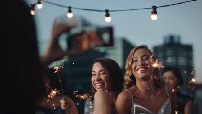 美丽的年轻女子跳舞与烟火女友庆祝新年前夜在迷人的派对穿着时尚时尚的朋友使用智能手机分享庆祝视频在社交