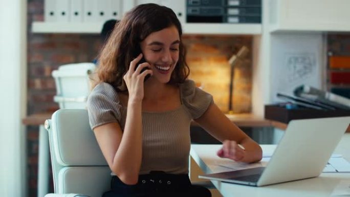 年轻的微笑女商人在办公桌前接听手机，以慢动作拍摄笔记本电脑
