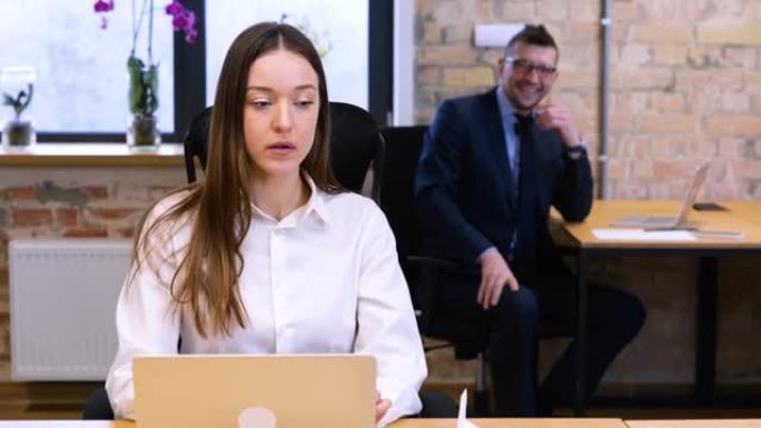 实时视频。两个白人同事男人和女人在办公室工作。女性正在笔记本电脑上打字，男性将纸飞机降落在桌子上。