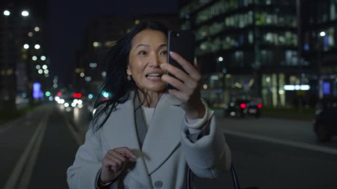 真实的亚洲中年女商人的肖像，晚上在城市街道上使用智能手机进行视频通话。出差中的女CEO，在网上与家人
