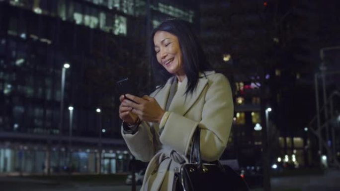 中年亚洲妇女的肖像晚上在城市街道上行走，使用智能手机。优雅的女商人微笑，发短信，在社交媒体上聊天，通