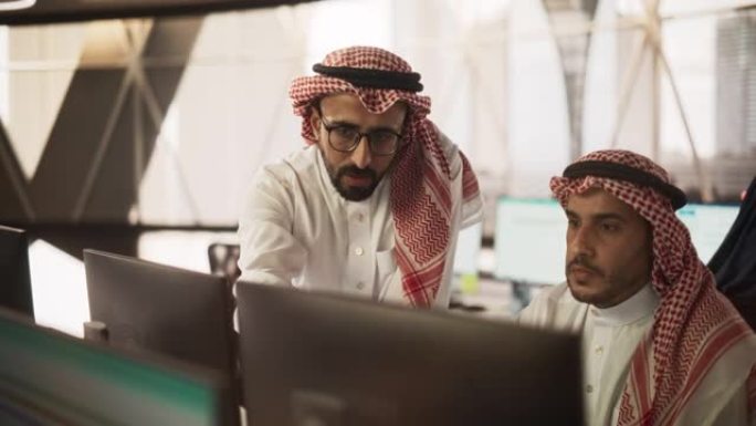 阿拉伯平面设计师从事计算机工作，与团队负责人讨论项目。中东同事在现代办公室工作，穿着传统的白色tho