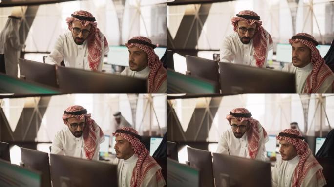 阿拉伯平面设计师从事计算机工作，与团队负责人讨论项目。中东同事在现代办公室工作，穿着传统的白色tho