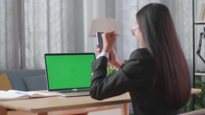 亚洲女商人穿着西装，绿屏电脑看着手机，在家工作时对项目投标结果感到满意。