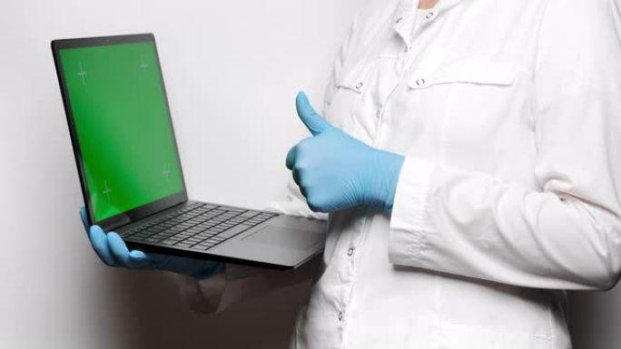 穿着白色外套和蓝色手套的医生拿着一台带有跟踪点的绿色屏幕的笔记本电脑，竖起大拇指。4k，复制空间，慢