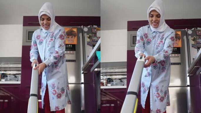 中年穆斯林妇女在厨房戴着头巾的无线吸尘器地板上吸尘。家庭主妇用电子设备清洁洗涤家庭。家电技术在日常生
