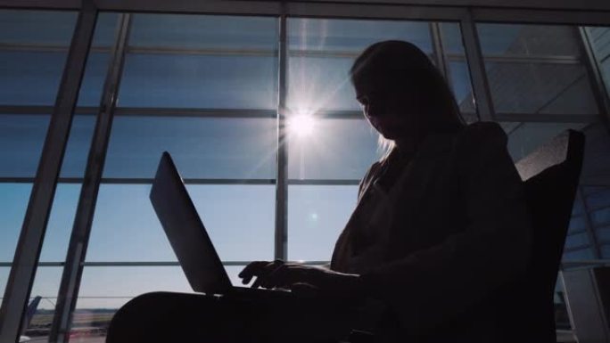 一名男子在机场航站楼的大窗户附近使用笔记本电脑的剪影