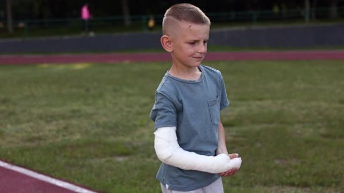悲伤的白人男孩断手，夏天在运动场上走在户外。四肢骨折和石膏骨折的9岁孩子。暑假骨折损伤。健康医疗理念