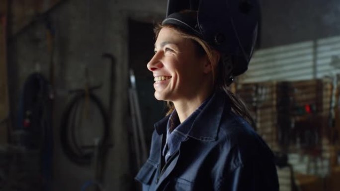 电影特写年轻的专业女焊工在制服工作服和手套脱下防护头盔和微笑。铁匠对她在车间里做金属加工后的成就感到