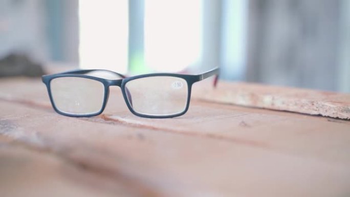 黑框大凸透镜眼镜在建筑工地模糊背景上的木板上特写镜头，平滑的相机移动。房间维修的准确性，工作时划伤的