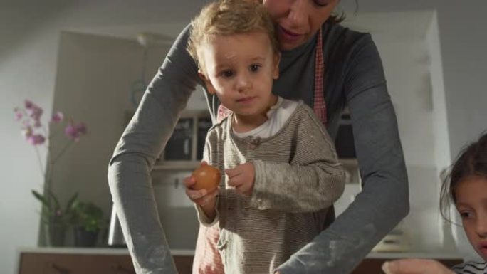 一个蹒跚学步的小男孩在母亲准备面团烘烤时轻轻地玩着鸡蛋的肖像。孩子们在厨房里帮助母亲，学习如何做饭和