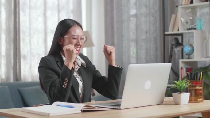 穿着西装的亚洲女商人看着电脑屏幕，在家工作时很高兴赢得项目投标