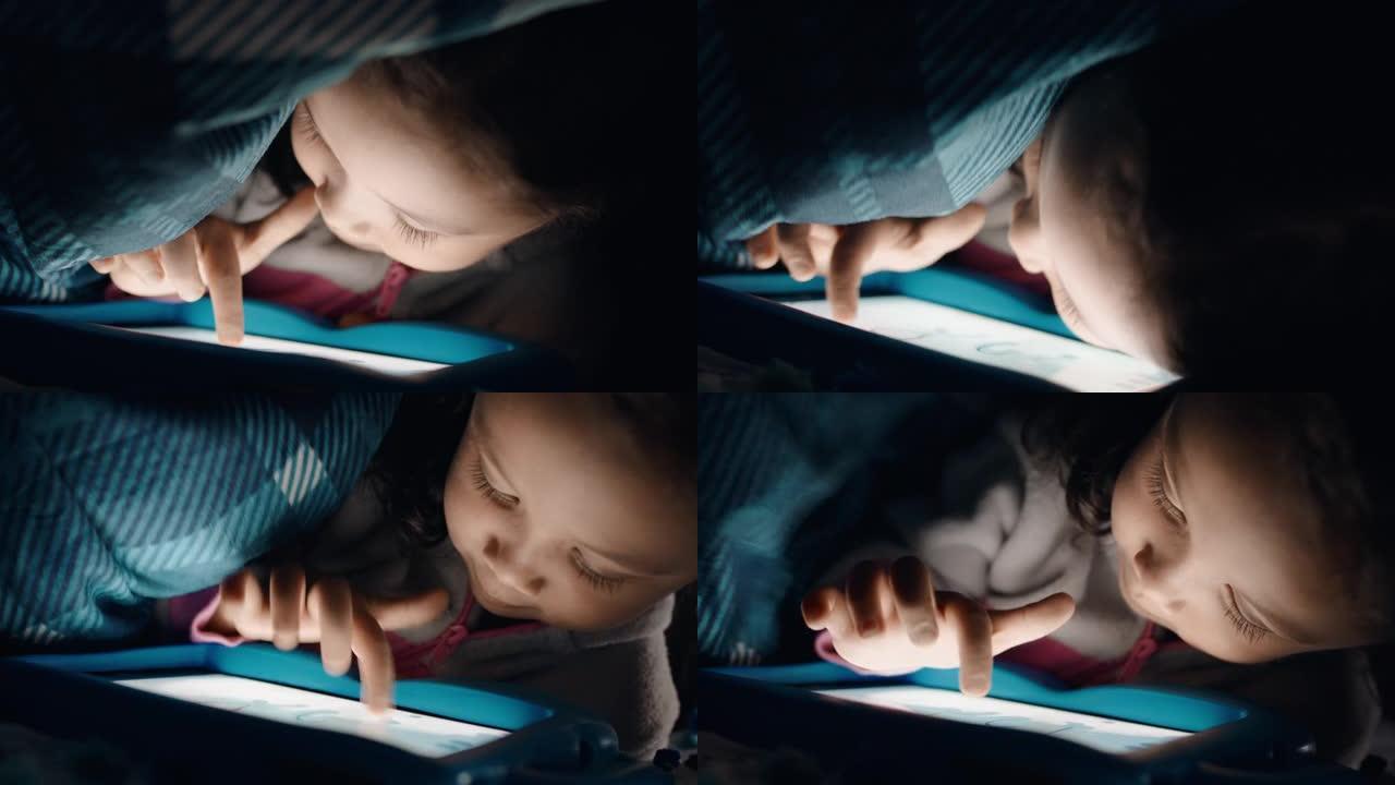 可爱的小女孩在毯子下使用数字平板电脑享受触摸屏技术玩游戏睡觉时玩得开心