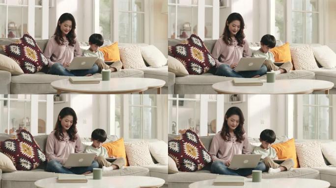 年轻的亚洲母亲在家工作，儿子在她身边玩电脑游戏