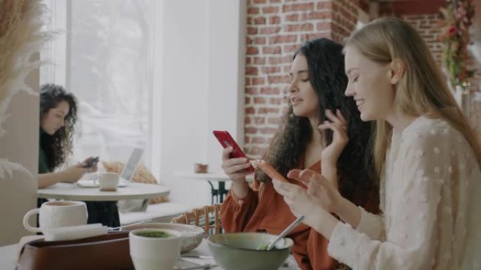 快乐的年轻女性看着智能手机屏幕聊天，坐在咖啡馆的桌子上分享内容
