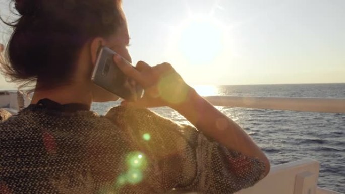 快乐的年轻女子在海上旅行的日出时微笑着在智能手机上聊天。在游轮度假渡轮上。