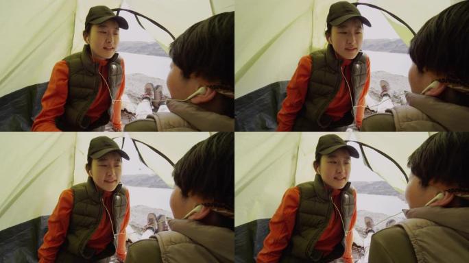 两个亚洲女性朋友坐在帐篷里，一个与另一个共享耳机