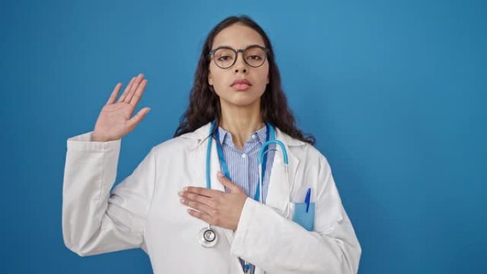 年轻美丽的西班牙裔女医生用手在孤立的蓝色背景上发誓