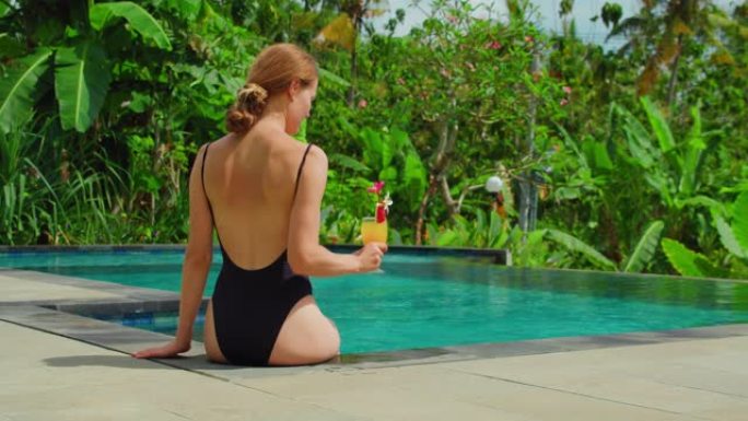 女人在游泳池边放松，手里拿着一杯橙汁鸡尾酒。背景中的热带棕榈树森林和水稻梯田。