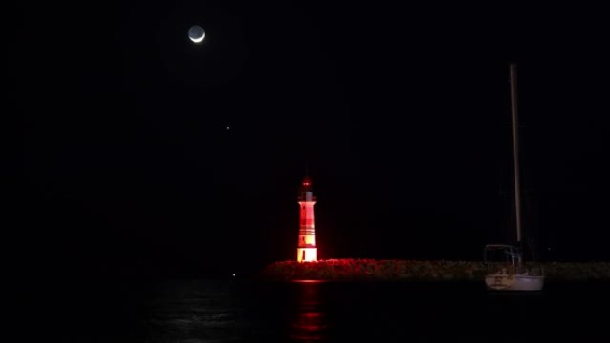 金星首先在红色灯塔上方落下，然后是月亮。新月穿过灯笼的中心。大海中一艘帆船的延时图像。