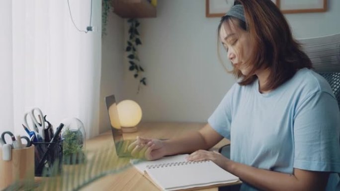 亚洲女人坐在餐桌旁，在笔记本上做笔记。用电脑笔记本电脑在桌子上工作，文件文书工作。