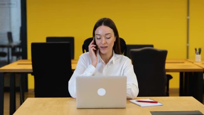 慢动作视频。白人妇女坐在她的工作台上，在电脑上工作，然后看着她的手机，开始说话。