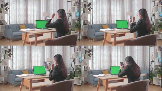 亚洲女商人穿着西装，绿屏电脑看着手机，在家工作时对项目投标结果感到满意。