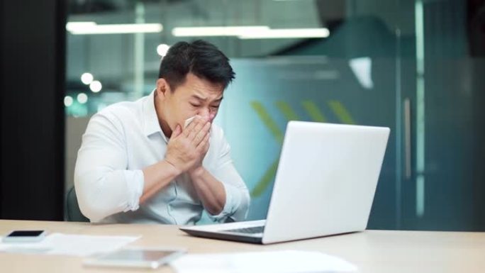 英俊的亚洲男性工人患有过敏，打喷嚏流鼻涕。员工坐在办公室里咳嗽感冒或流感
