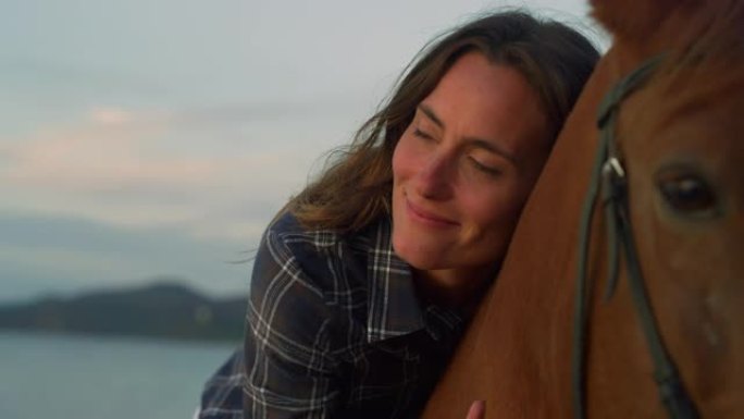 日落时在海边海滩上骑马的美女的特写肖像。职业女性马术在看海的同时拥抱和爱抚着她的纯种马。自由概念