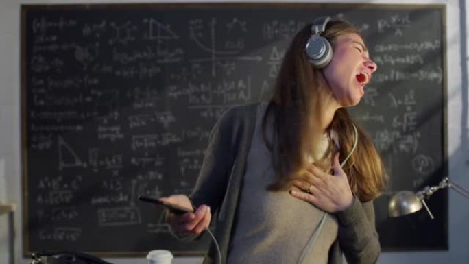 年轻快乐的女老师或学生的肖像，戴着耳机，用智能手机听音乐，在大学或大学教室里用公式在黑板前跳舞