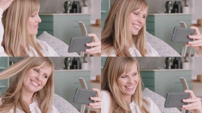 美丽的金发女人在家中使用智能手机进行视频聊天，炫耀发型与朋友分享生活方式，享受在线交流