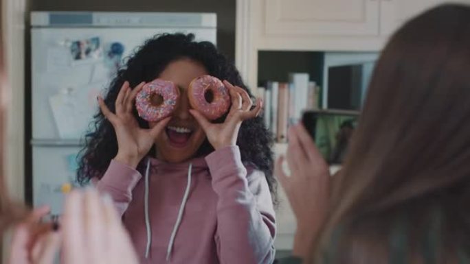 有趣的十几岁的女孩与甜甜圈合影，在社交媒体上分享智能手机，享受周末在厨房闲逛的乐趣