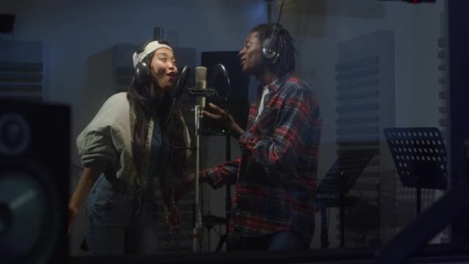 录音棚里两位多民族职业歌手的肖像，戴着耳机，对着麦克风热情地唱歌。年轻的黑人和亚洲妇女合作演唱了有关