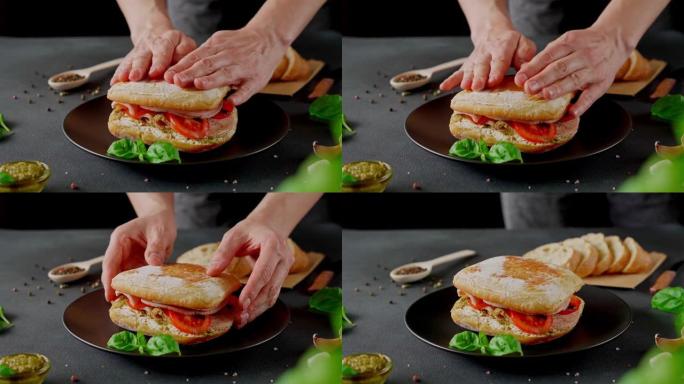 男人的手准备了Chiabatta三明治，上面有火腿，罗勒，香蒜酱，西红柿在黑色背景上。食物背景