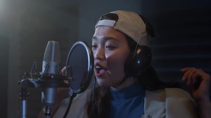 用耳机在专业录音室录制专辑的年轻亚洲女歌手的特写肖像。美丽的富有表现力的女人带着情感唱歌，开始了成功