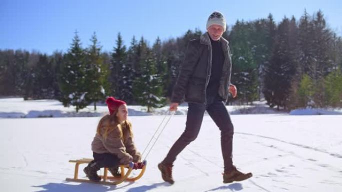 SLO MO TS祖父在阳光下将他的女儿拉到雪橇上，穿过冰冻的湖面