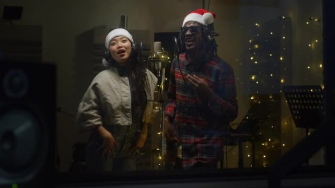 在录音棚里用圣诞老人帽子和耳机录制的两位不同歌手的特写肖像。年轻的黑人男性和亚洲女性艺术家在背景中与