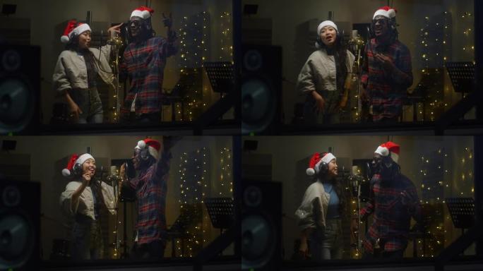在录音棚里用圣诞老人帽子和耳机录制的两位不同歌手的特写肖像。年轻的黑人男性和亚洲女性艺术家在背景中与