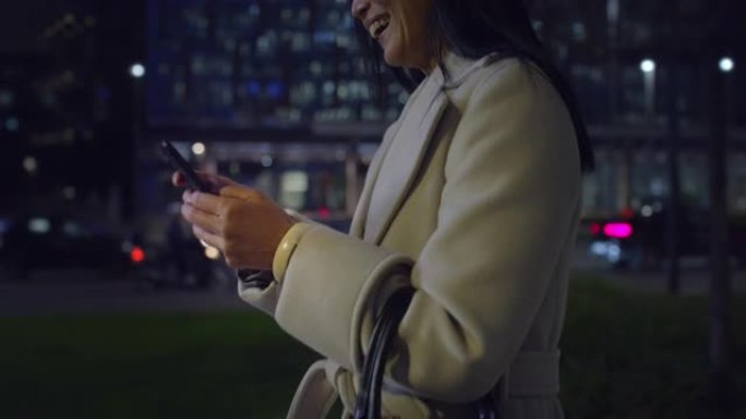 用智能手机在夜间走在城市街道上的中年亚洲妇女的手中关闭。优雅的女商人微笑，发短信，在社交媒体上聊天，
