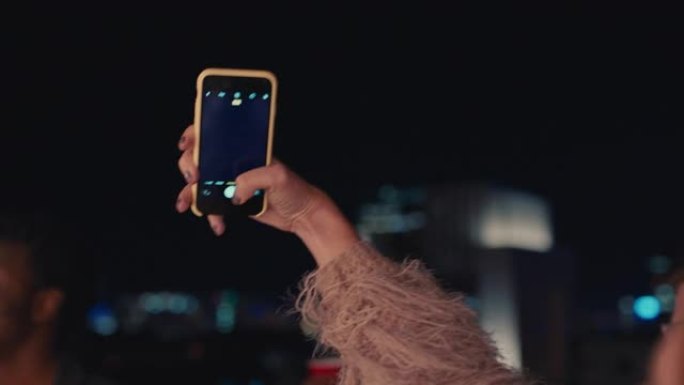 一群多种族的朋友晚上在屋顶派对上跳舞，美丽的年轻女子使用智能手机在社交媒体上分享狂野的周末生活方式拍