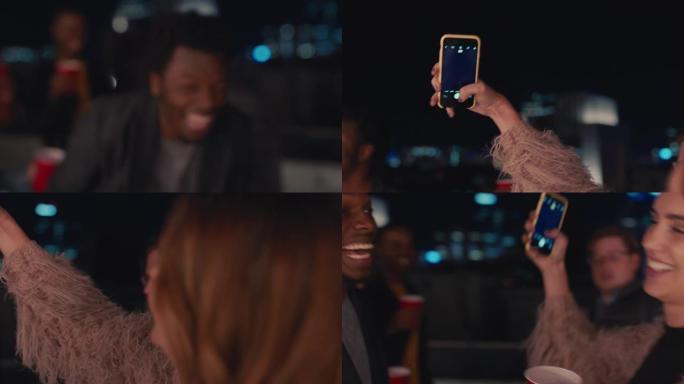 一群多种族的朋友晚上在屋顶派对上跳舞，美丽的年轻女子使用智能手机在社交媒体上分享狂野的周末生活方式拍